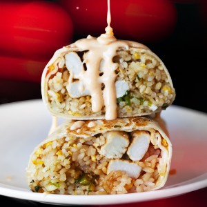 A burrito cut in half, filled with fried rice and chicken, topped with creamy sauce being dribbled on top.