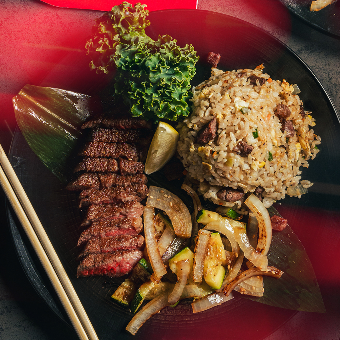 Sliced steak, fried rice, sautéed onions and zucchini, plus greens arranged on a black plate.