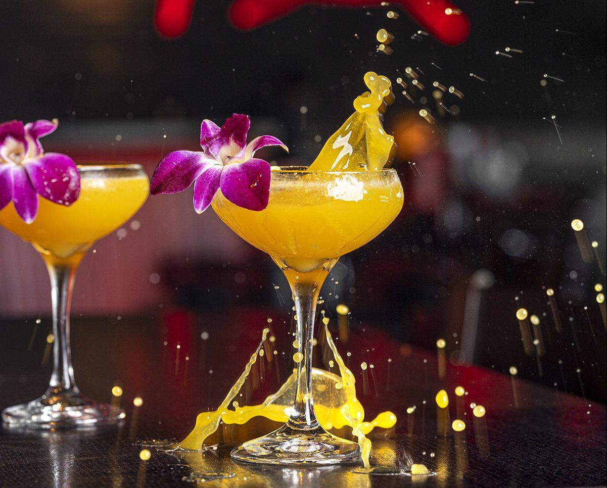 A bright yellow cocktail in a coupe glass garnished with a purple orchid, mid splash, with droplets and liquid scattering across a dark table. Another similar cocktail is in the background.