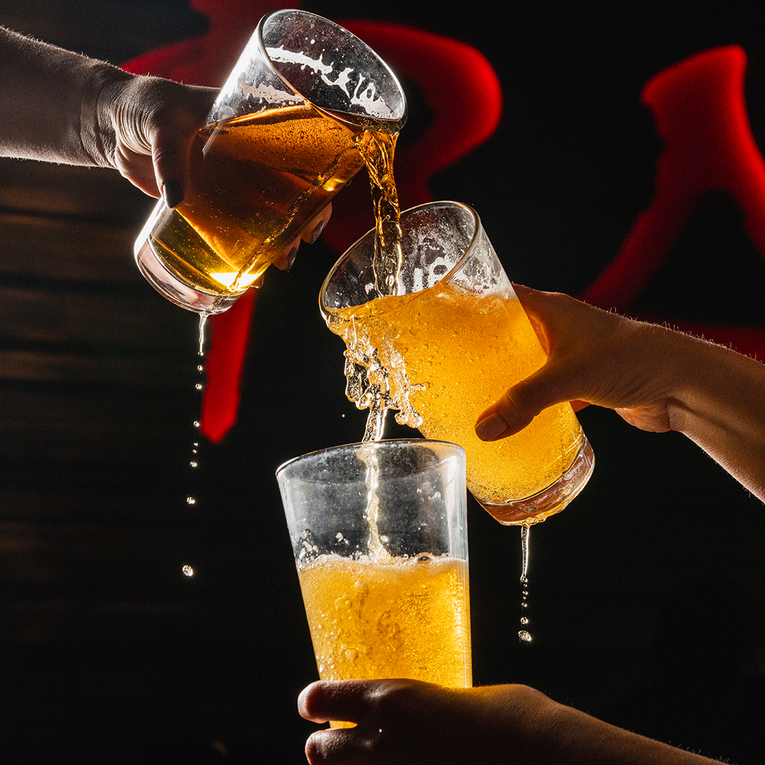Three hands clinking and pouring beer into glasses, with beer splashing and dripping, against a dark background with red light accents. Glasses are in a vertical line.