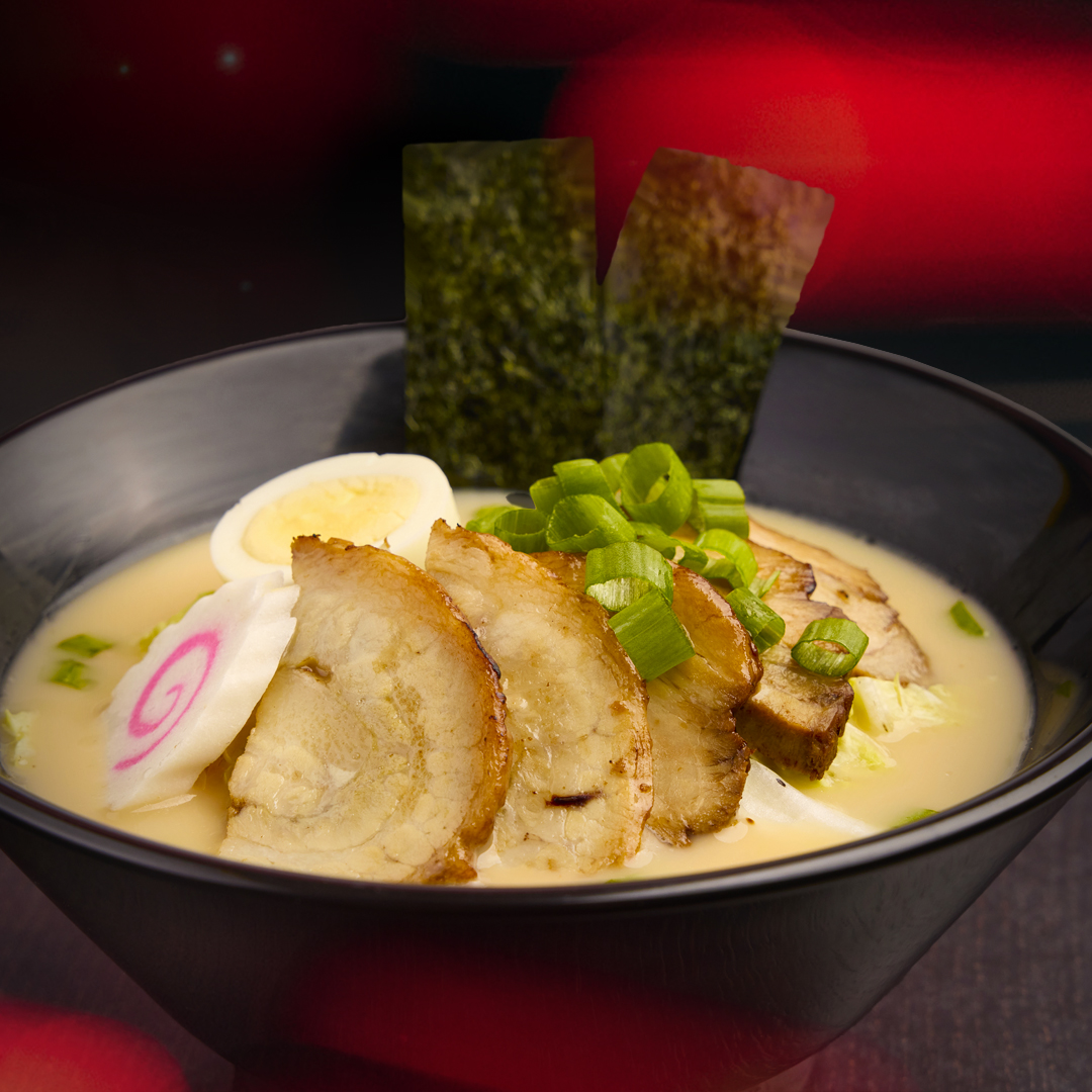 A bowl of chashu ramen topped with slices of braised pork, a marinated egg, fish cake with a pink swirl, green onions, and two sheets of nori in a creamy broth.