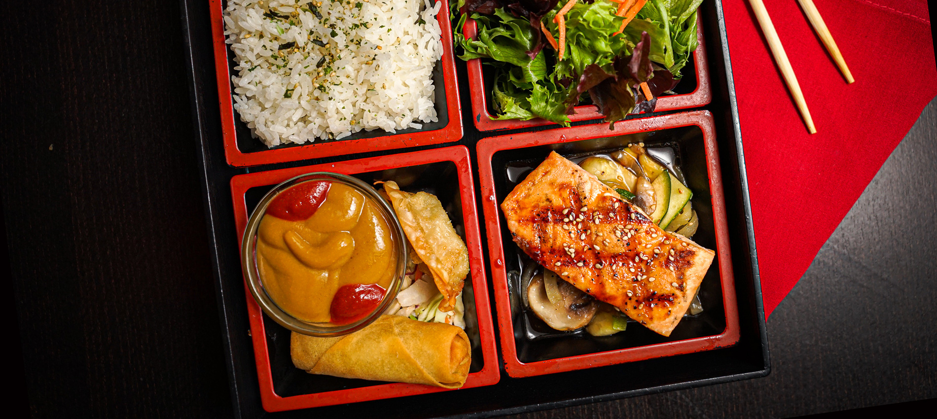 A close up of a bento box on a black table with grilled salmon, white rice, salad, spring roll, and dipping sauce. There is a red napkin underneath and chopsticks to the side.