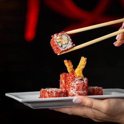 A hand holds chopsticks picking up a piece of tempura shrimp sushi roll coated in red seasoning from a white plate.