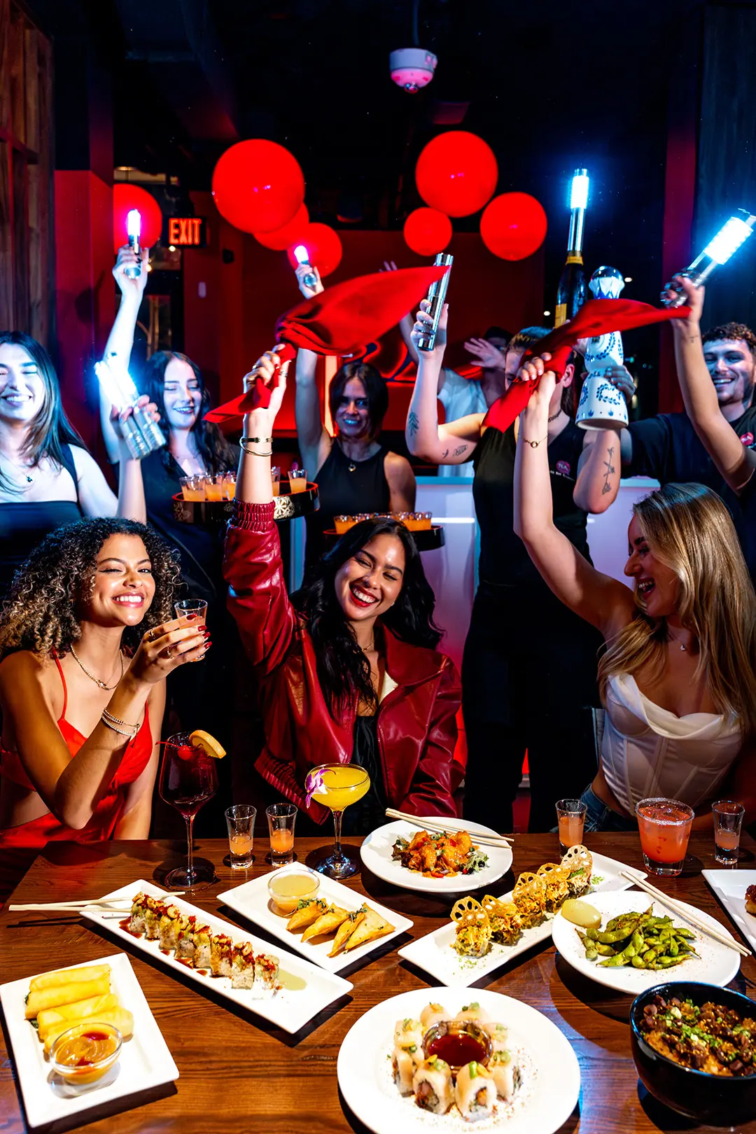 A lively group of friends celebrate at restaurant table filled with sushi rolls, appetizers, and cocktails, while servers in the background wave red napkins and hold glowing lights under vibrant red lanterns.