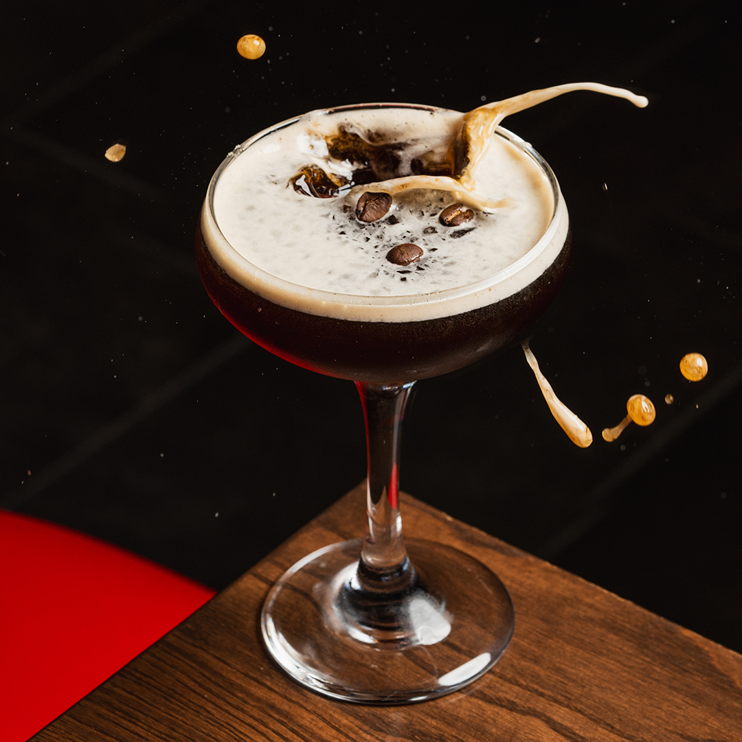 A close up of an espresso martini cocktail in a coupe glass, garnished with coffee beans, as a splash erupts from the creamy foam top against a dark background.