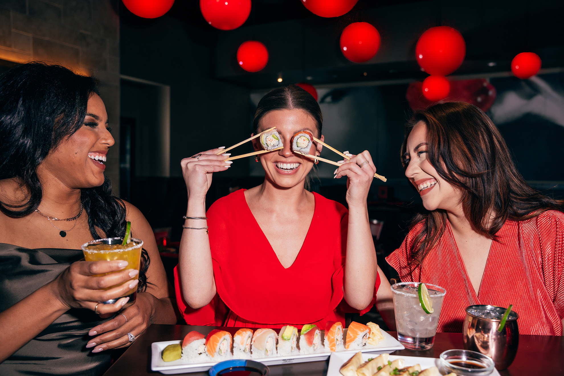 Three women enjoy drinks and a platter of assorted sushi rolls at a restaurant with red hanging lights. Woman in centre places 2 sushi rolls over her eyes and the three women laugh.