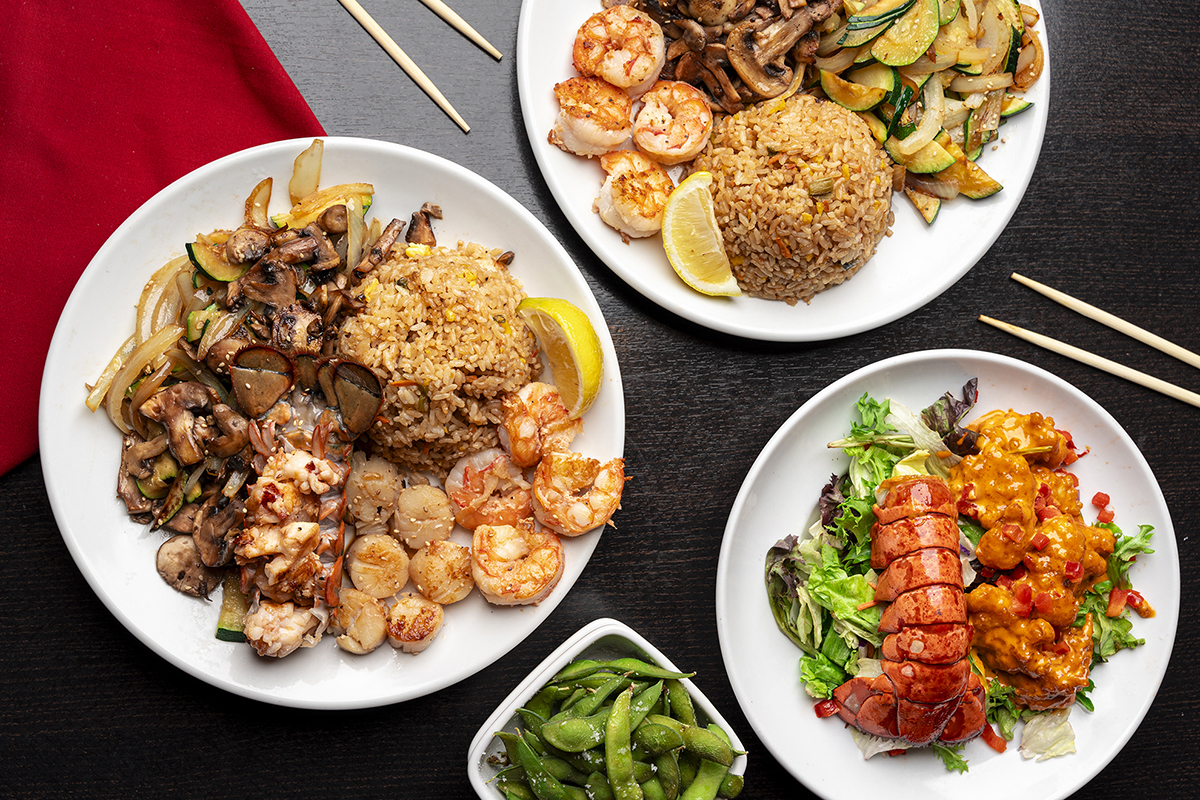 An overhead view of assorted entrées including grilled lobster tail, shrimp, scallops, sautéed mushrooms and vegetables, fried rice, spicy seafood over mixed greens, and a bowl of edamame, all arranged on white plates with chopsticks and a red napkin on a black wooden table.
