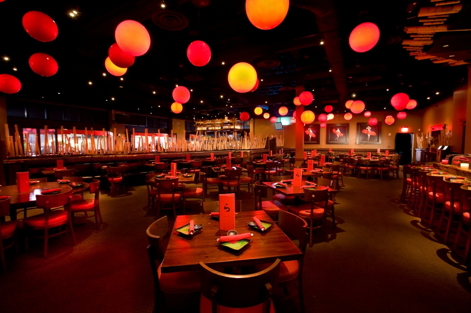 Interior shot with red accent lighting throughout. Dark ceiling with globe pendant lighting in a red, yellow and orange assortment. Wooden square tables are set with cutlery, napkins and menus