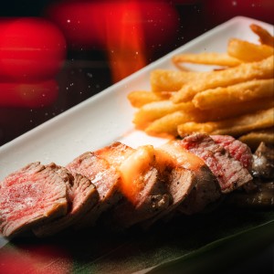 A close up of a rectangular white plate with slices of rare seared steak topped with melted butter, served alongside a portion of golden frites. Blurred red and black background.