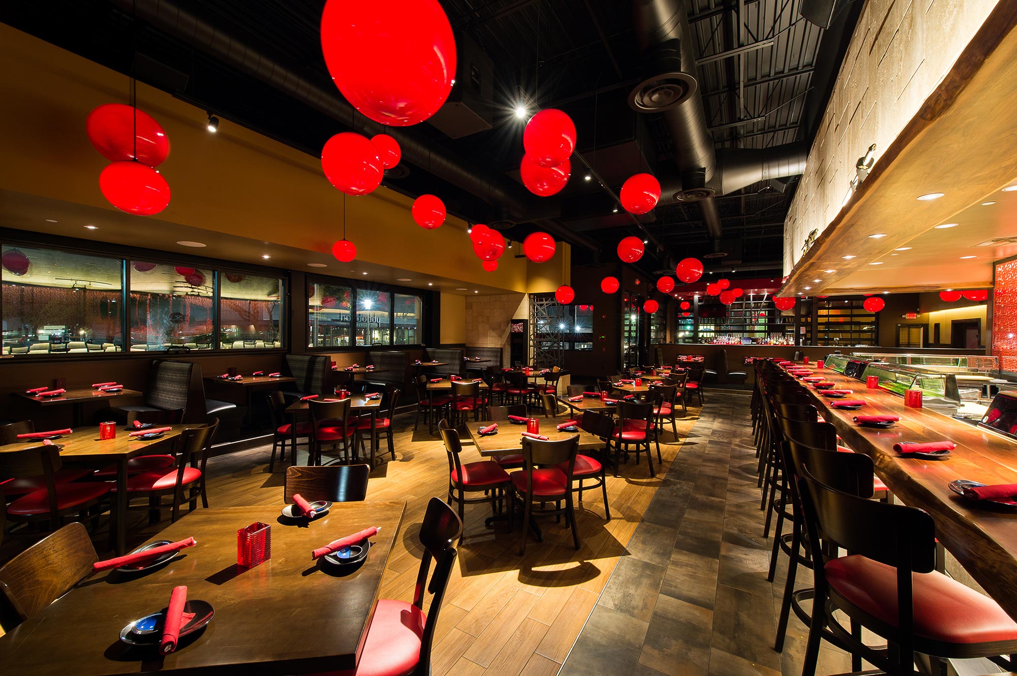 A restaurant interior features large red lights dotted around the ceiling. With wooden tables with red napkins, and a long sushi bar with counter seating.