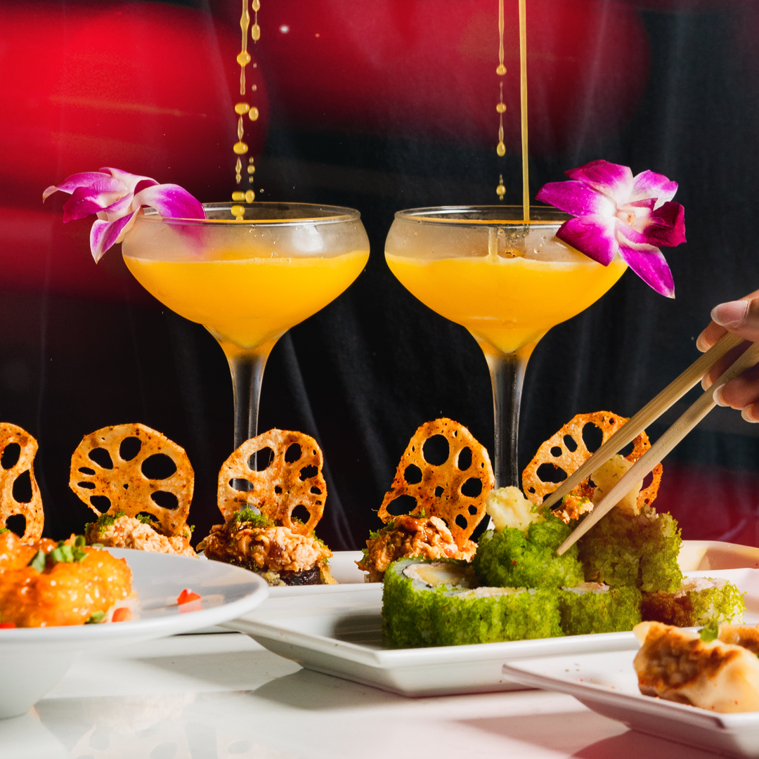 Two yellow cocktails garnished with purple orchids are served alongside sushi rolls coated in green flakes, crispy lotus chips, and spicy seafood appetizers.