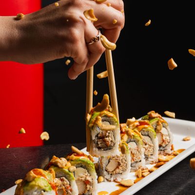 A hand uses chopsticks to pick up a sushi roll topped with avocado, sauce, and crushed peanuts, with more rolls and scattered peanuts on a rectangular plate.