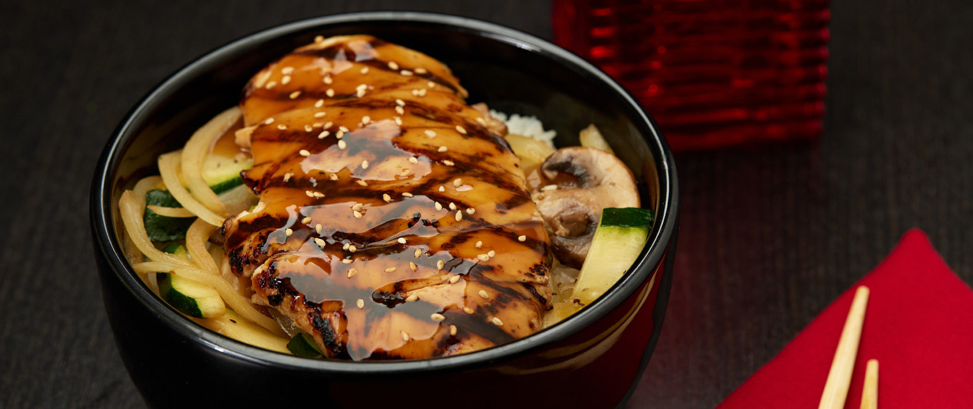 A bowl of grilled chicken glazed with teriyaki sauce, topped with sesame seeds, served over sautéed vegetables and white rice, with chopsticks and a red napkin on the side.