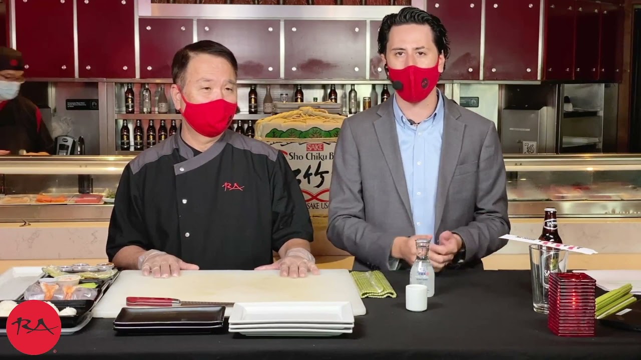 Two people stand behind a counter in a sushi restaurant, one wearing a chef's jacket with the RA Sushi logo and the other in a suit. Sushi ingredients, plates, chopsticks, and bottles are arranged in front of them.