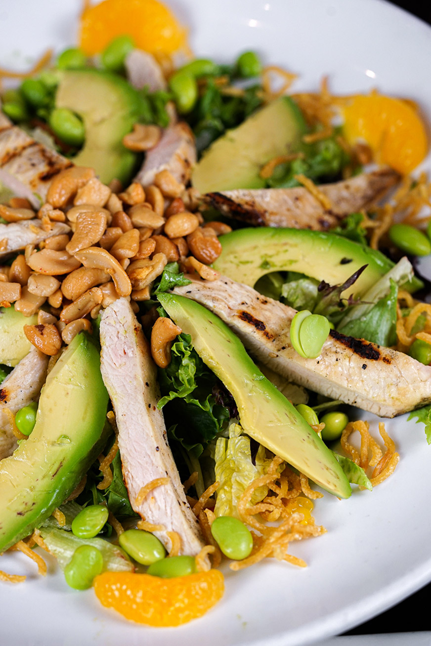 A close up of Nutty Grilled Chicken Salad featuring sliced grilled chicken breast, avocado, mandarin orange segments, edamame, mixed greens, crispy wonton strips, and a generous topping of roasted peanuts.