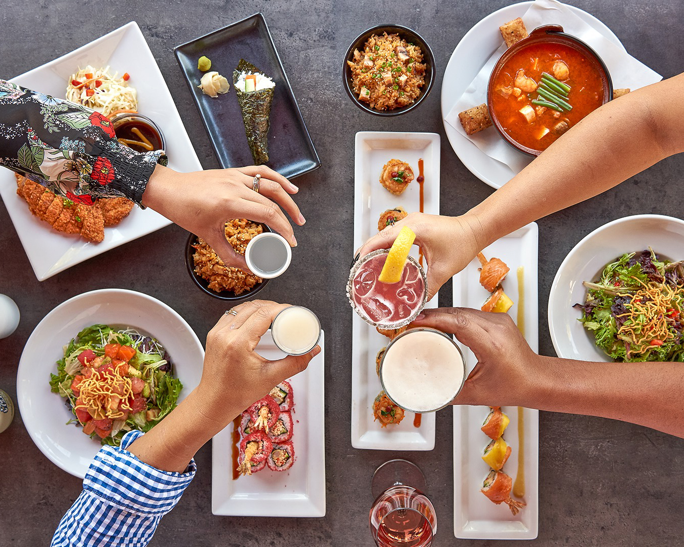 Four people raise drinks over a table set with sushi rolls, seaweed hand rolls, salad, shrimp soup, fried rice, and crispy breaded chicken.